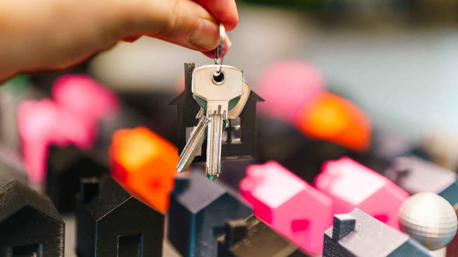 Hand holding house keys next to colorful miniature houses